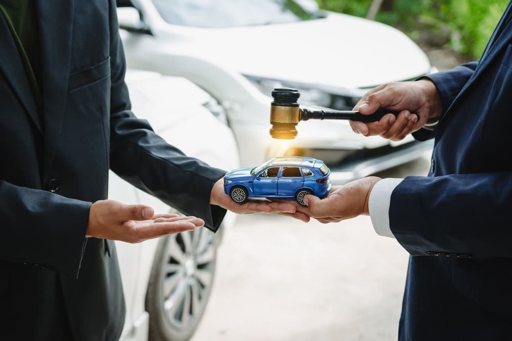 Two Asian individuals meet with an insurance agent in front of a damaged vehicle to discuss a car insurance claim. They review documents, go over policy details, and address key concerns.