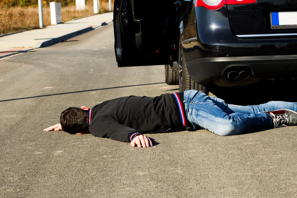 A man lies on the road behind a black car after a collision. The pedestrian is on the ground following the accident.