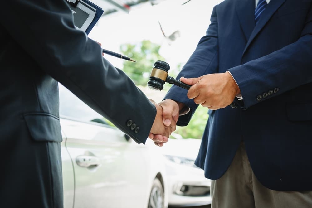 Two Asian individuals meet with an insurance agent in front of a damaged vehicle, reviewing documents, discussing policy details, and addressing crash and repair costs.