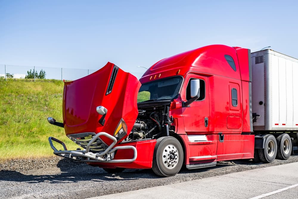 A red semi-truck with a dry van trailer sits disabled on the road shoulder, hood open, awaiting a tow truck or roadside assistance for repairs after an accident.